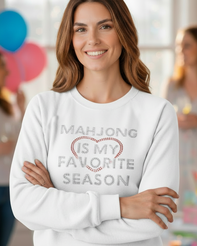 Woman wearing a white sweatshirt with 'Mahjong is my favorite season' text, standing in a room with blurred people in the background.