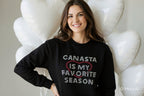 Woman wearing a black sweatshirt with 'CANASTA IS MY FAVORITE SEASON' text, standing against a white balloon background.