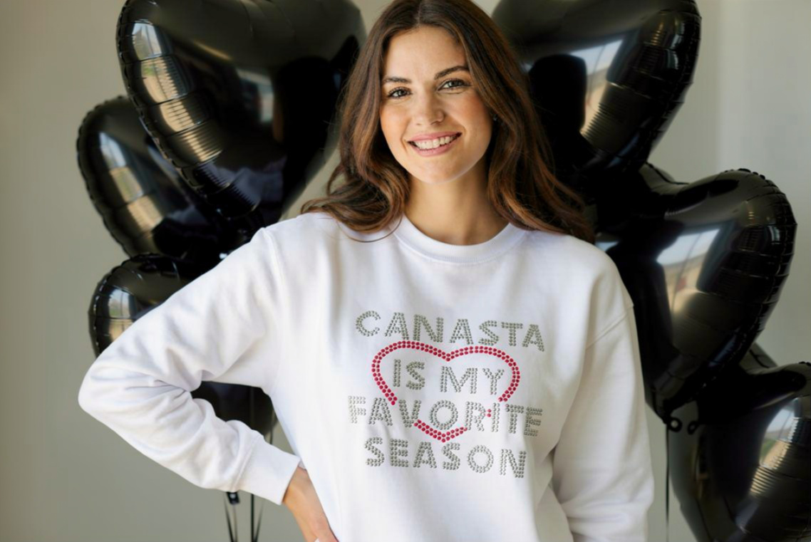 Woman wearing a white sweatshirt with 'Canasta is my favorite season' text, standing in front of black balloons.