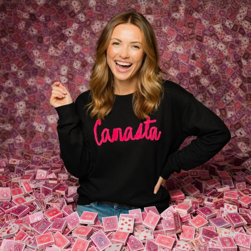 Woman wearing a black sweatshirt with 'Canasta' in pink, surrounded by playing cards against a patterned background.