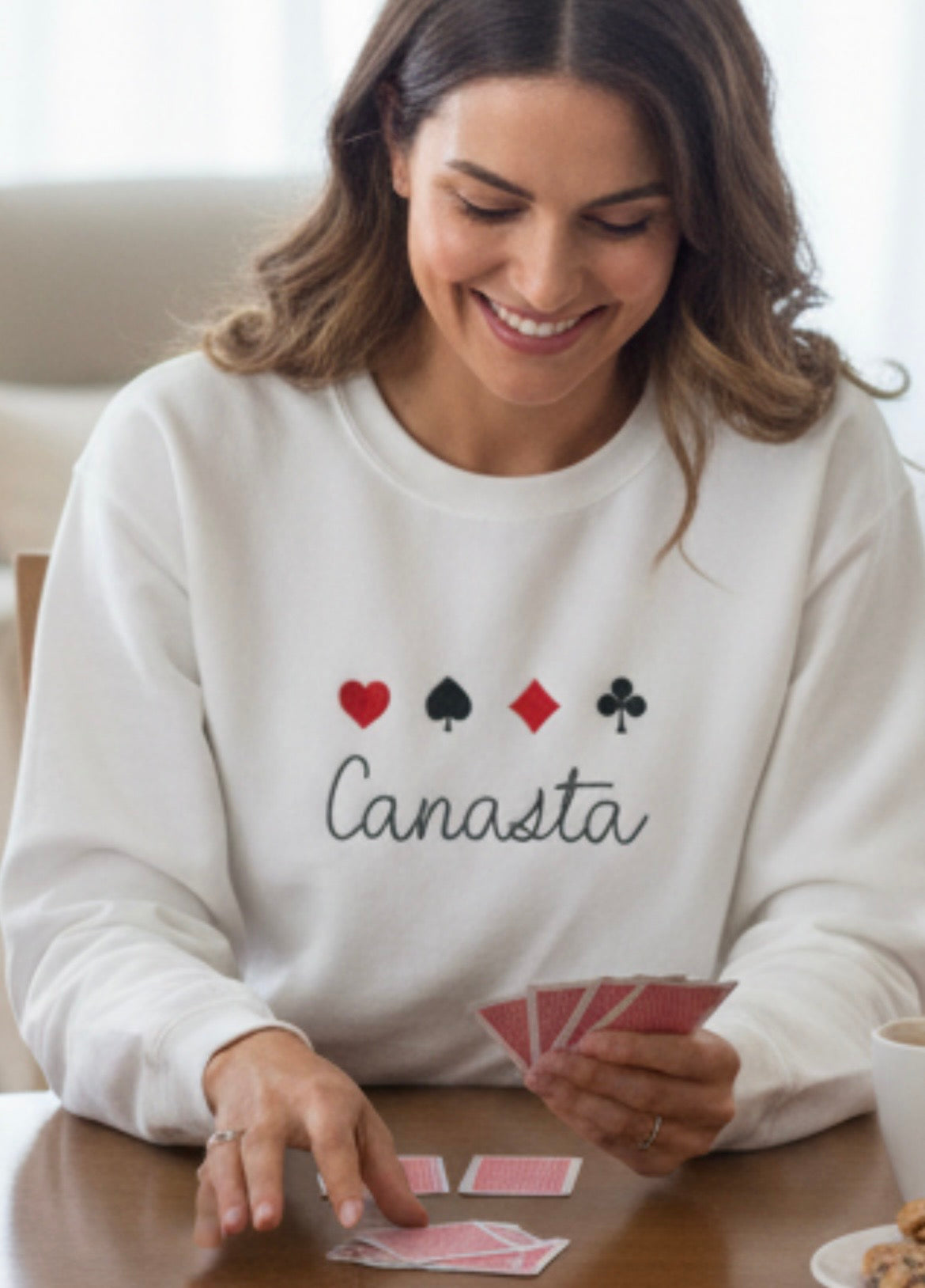 Woman wearing a white sweatshirt with 'Canasta' and card symbols, holding playing cards.