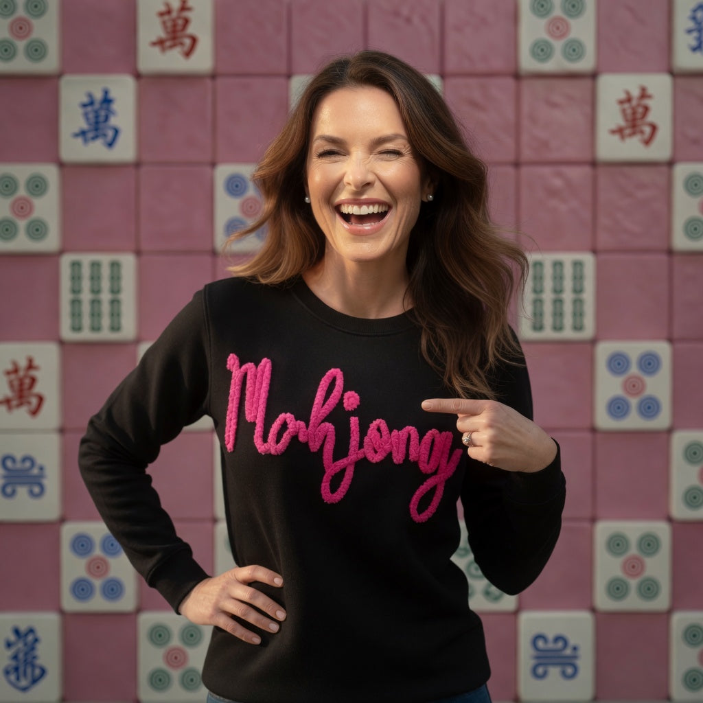 Woman wearing a black sweater with 'Mahjong' in pink text, pointing at it, against a pink tile wall with Mahjong tiles.