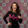Woman wearing a black sweater with 'Mahjong' in pink text, pointing at it, against a pink tile wall with Mahjong tiles.