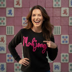 Woman wearing a black sweater with 'Mahjong' in pink text, pointing at it, against a pink tile wall with Mahjong tiles.