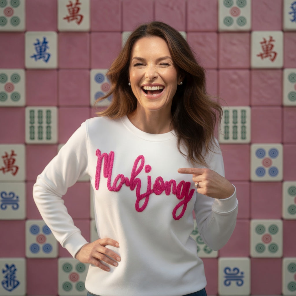 Woman wearing a white sweater with 'Mahjong' in pink text, pointing to it, against a pink tiled wall with Mahjong tiles.