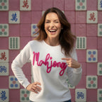 Woman wearing a white sweater with 'Mahjong' in pink text, pointing to it, against a pink tiled wall with Mahjong tiles.