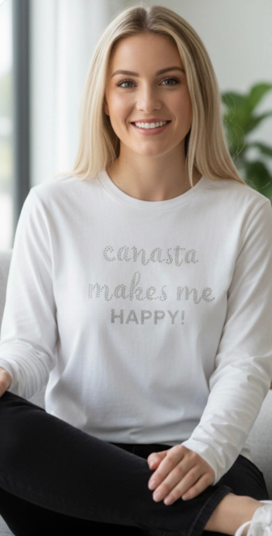 Woman wearing a white sweatshirt with 'canasta makes me happy!' text, sitting indoors.